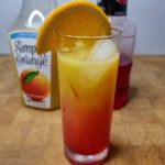 Sunrise mocktail with an orange slide in the glass and ingredient bottles behind the glass.