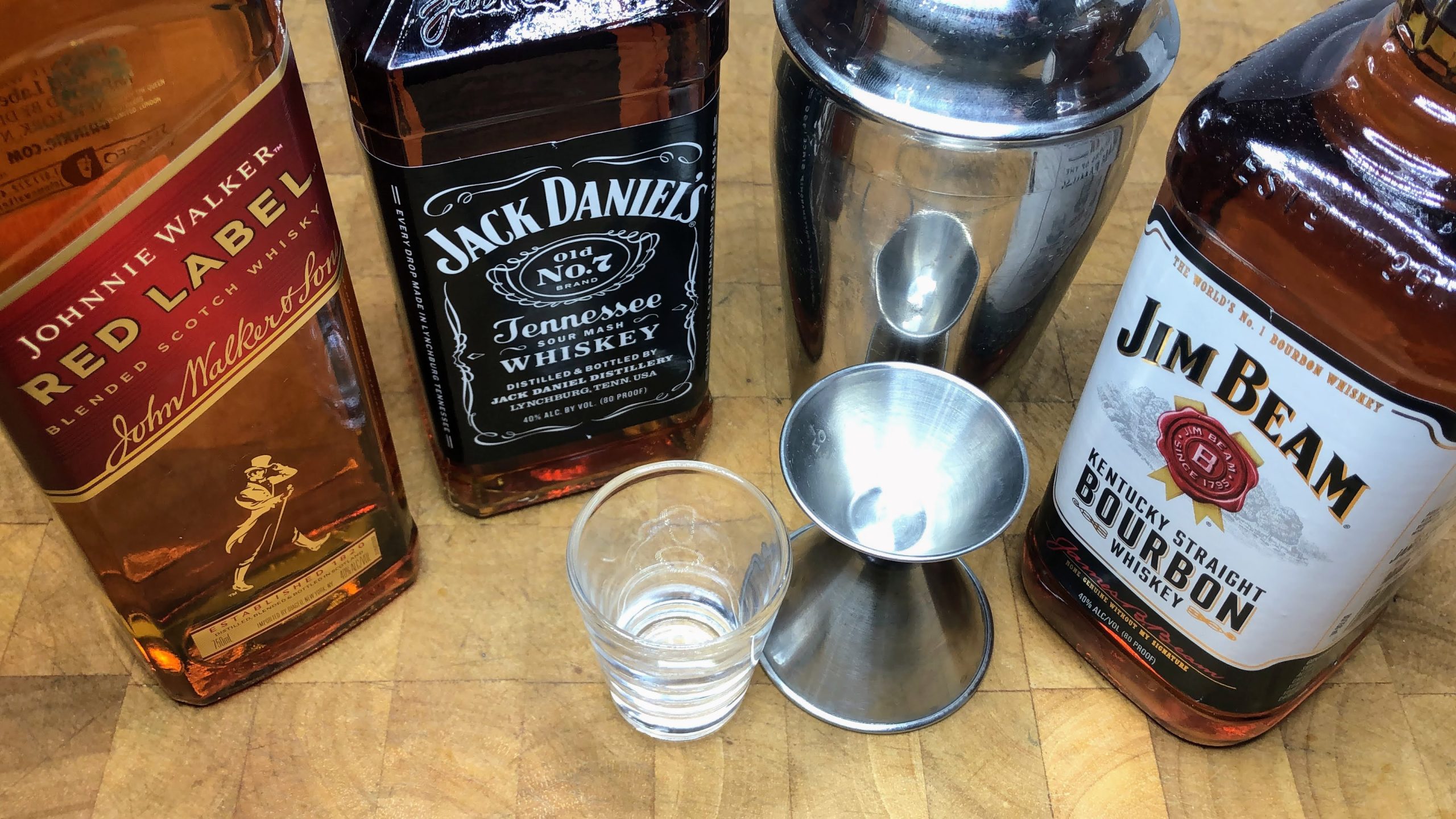 Bottles of Johnnie Walker, Jack Daniels and Jim Beam next to a shaker, empty shot glass and jigger.
