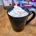 Vodka hot chocolate topped with whipped cream with ingredients behind the mug.