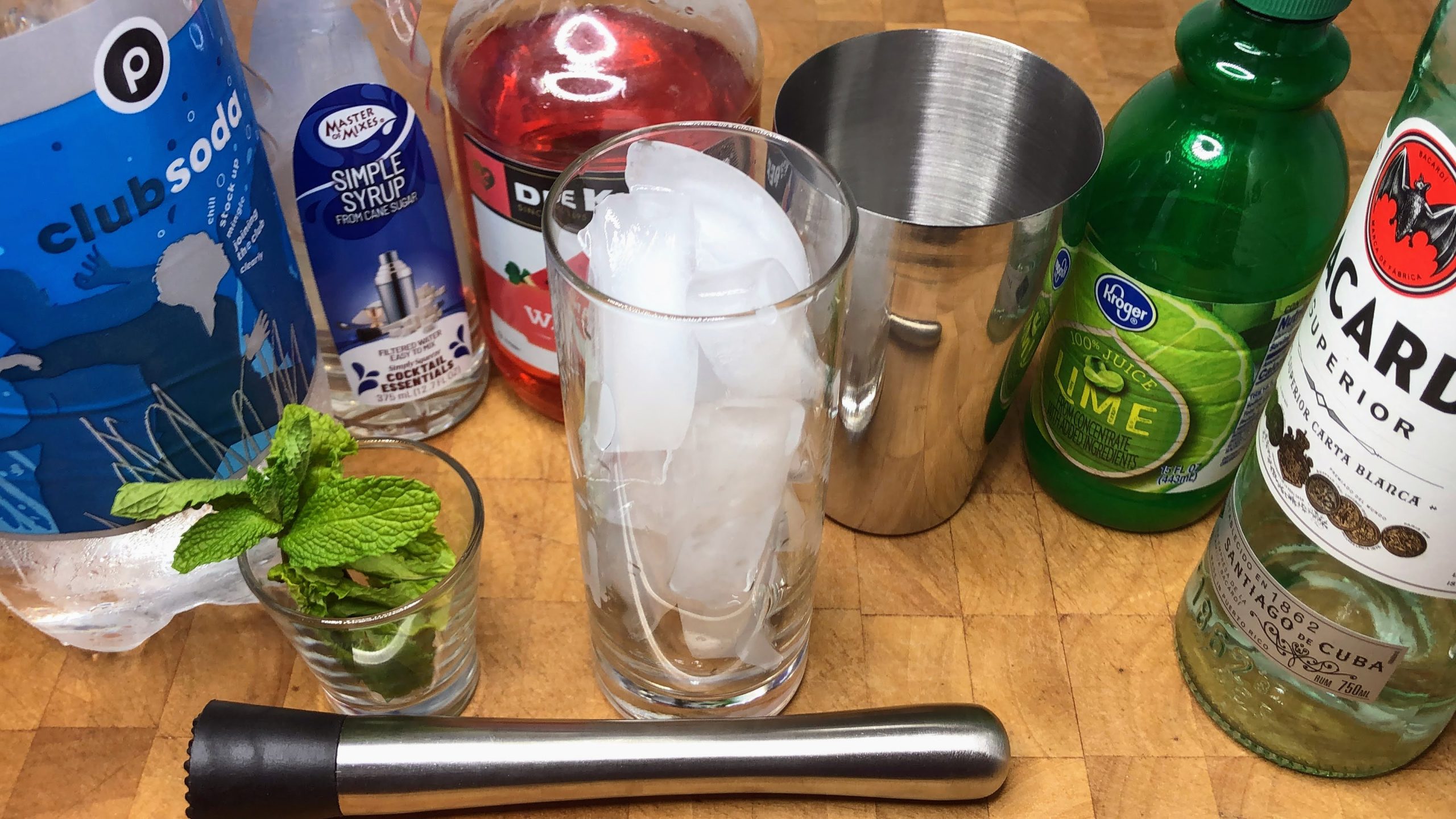 Bottles of club soda, simple syrup, watermelon pucker, lime juice and white rum next to mint, muddler, shaker and highball glass.