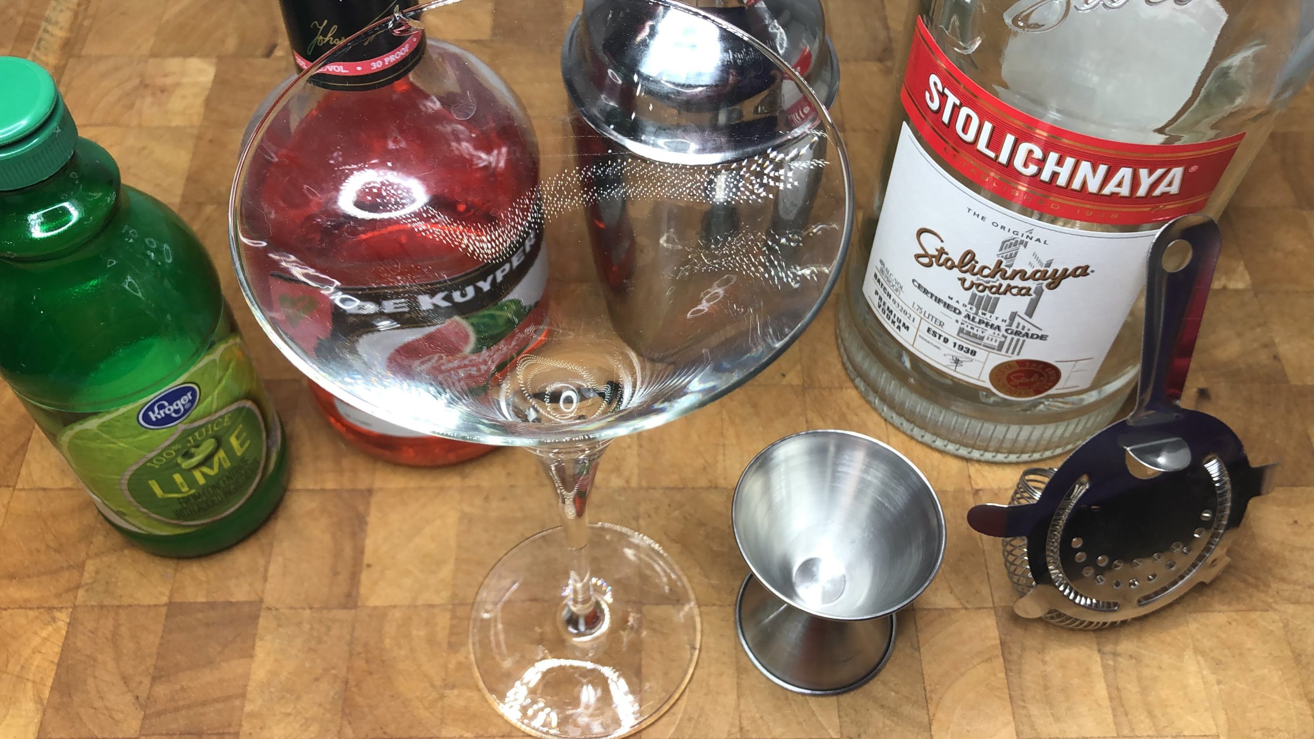 Martini glass, jigger, shaker and hawthorne strainer next to bottles of vodka, watermelon pucker and lime juice.