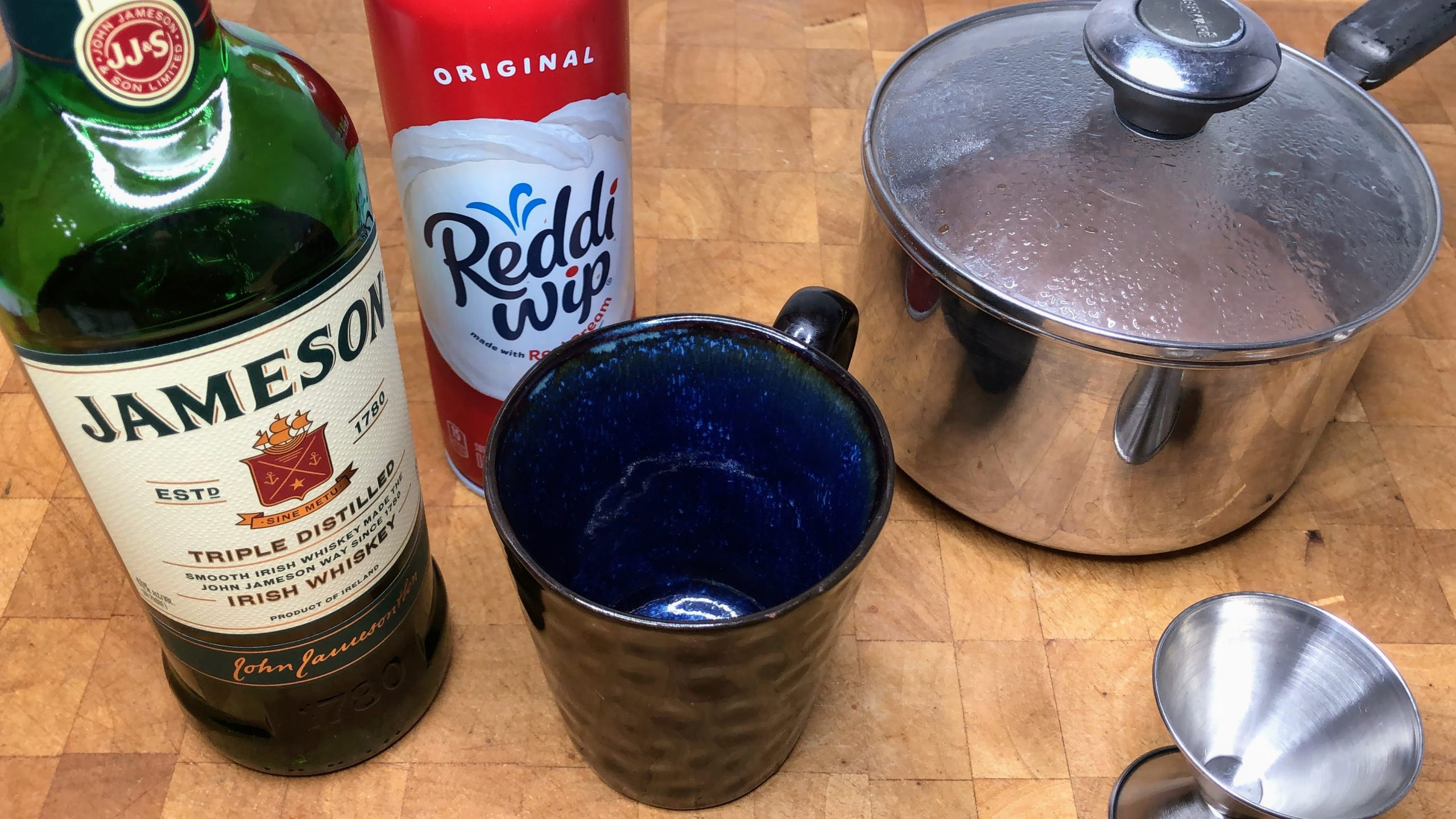 Pot of hot chocolate next to jigger, mug, whipped cream and irish whiskey.