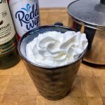 Whiskey hot chocolate topped with whipped cream in a mug with ingredients next to the mug.