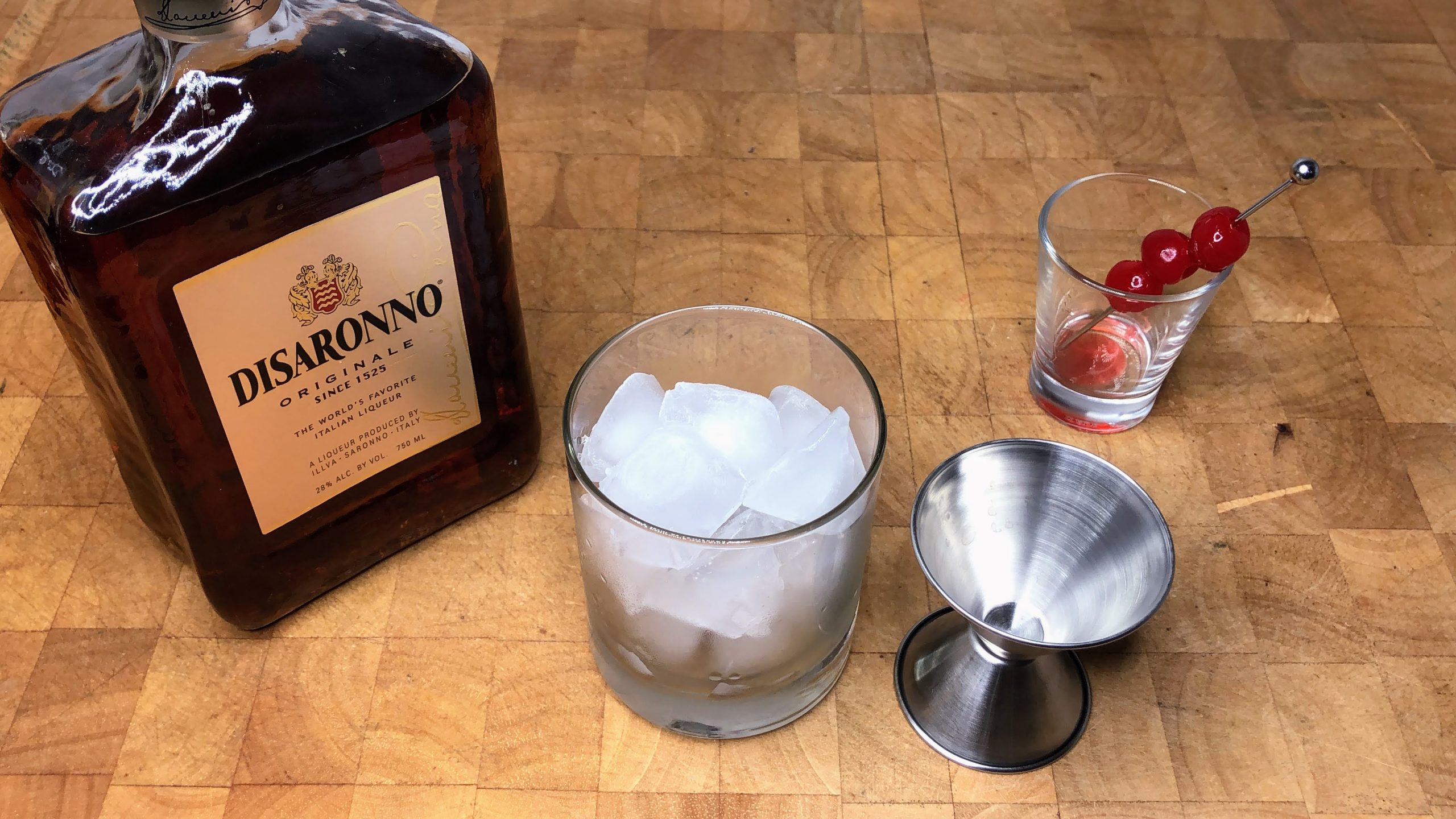 Rocks glass with ice next to shot glass with maraschino cherries, a jigger and a bottle of Amaretto.