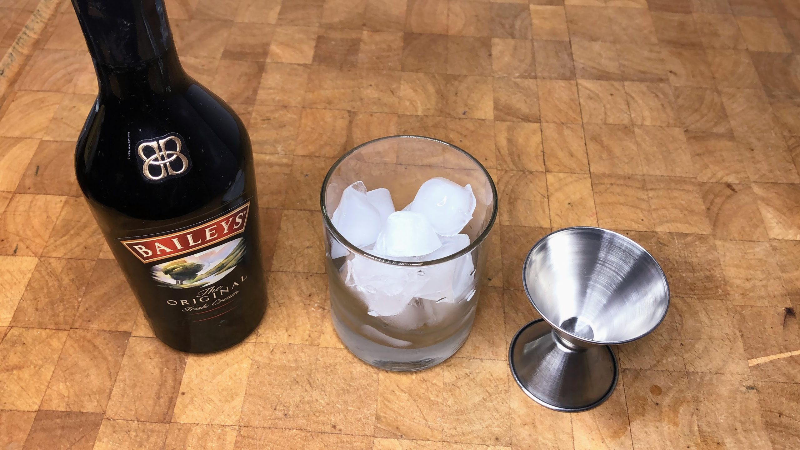 Bottle of baileys next to a rocks glass with ice and a jigger.
