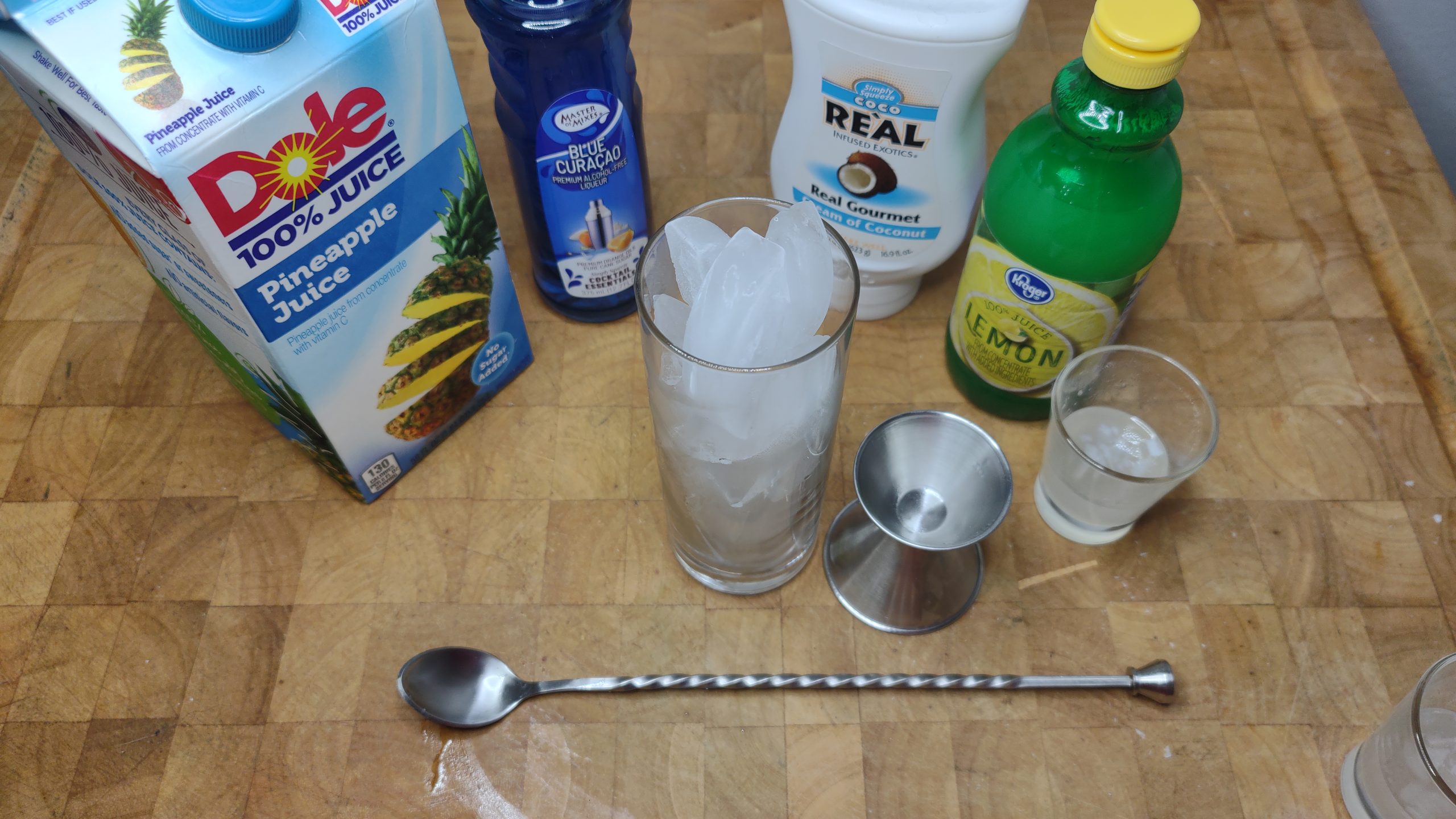Bar spoon, highball glass and jigger next to coconut simple syrup, lemon juice, cream of coconut, blue curacao and pineapple juice.