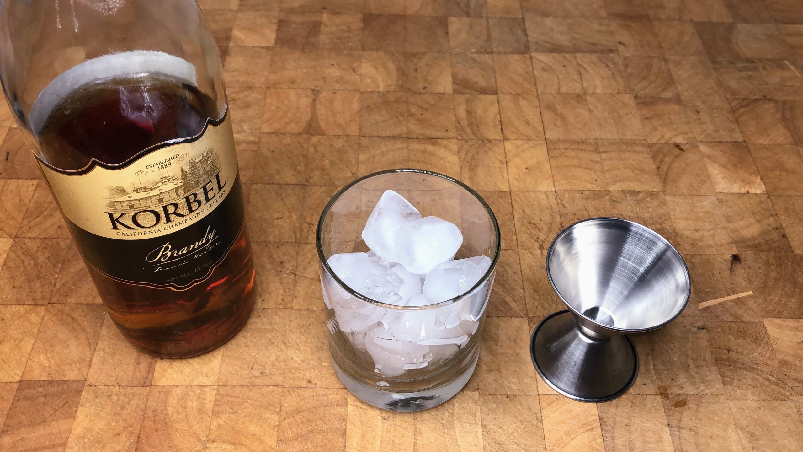 Brandy next to a rocks glass with ice and a jigger on a wooden table.