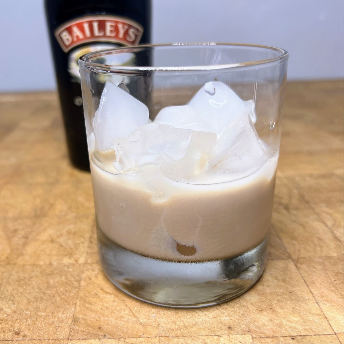 Bailey's on the rocks in a glass on a wooden table with a bottle of baileys in the background.
