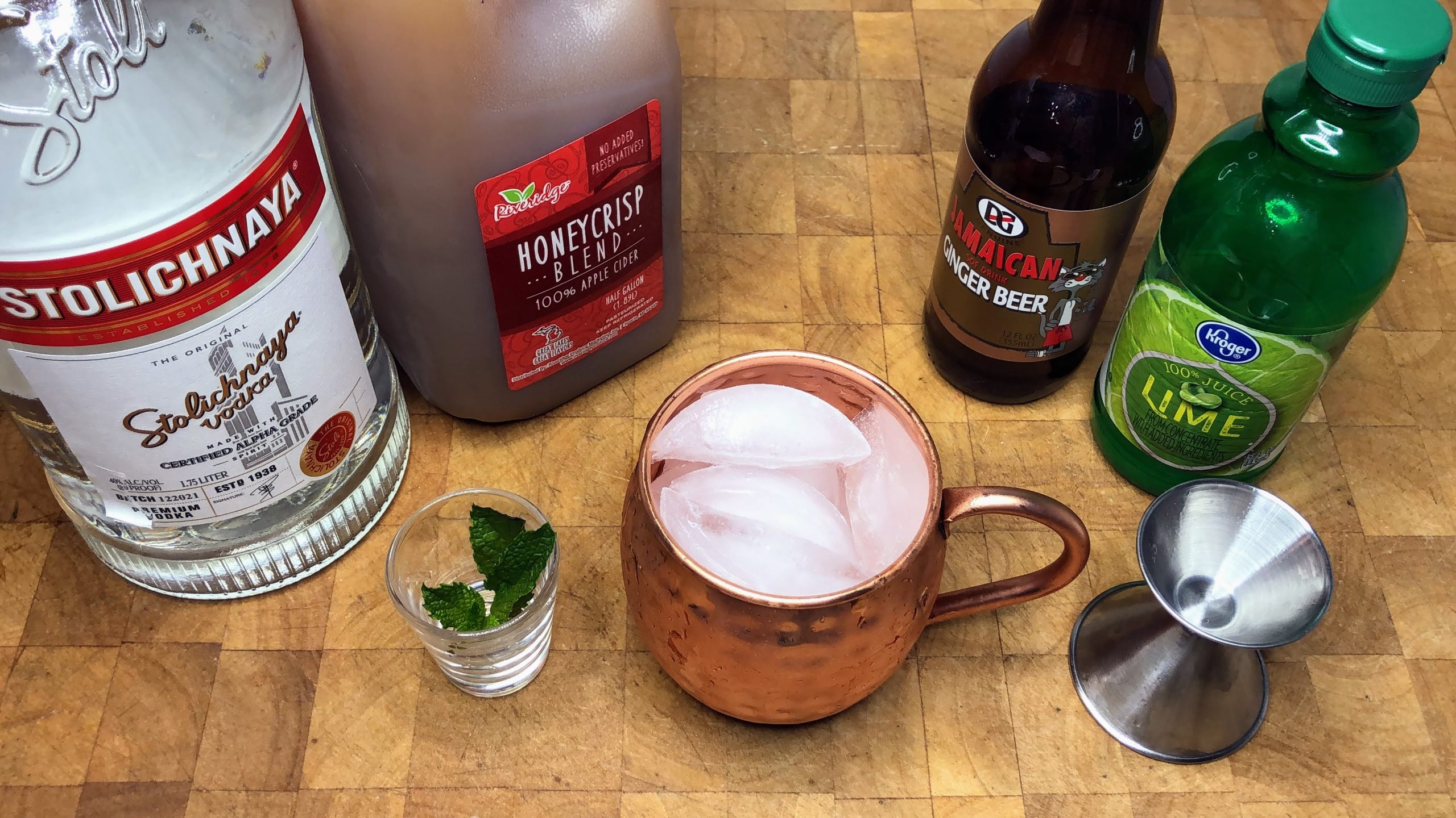 Bottles of vodka, apple cider, ginger beer and lime juice behind mint leaves, a copper mug with ice and a jigger.