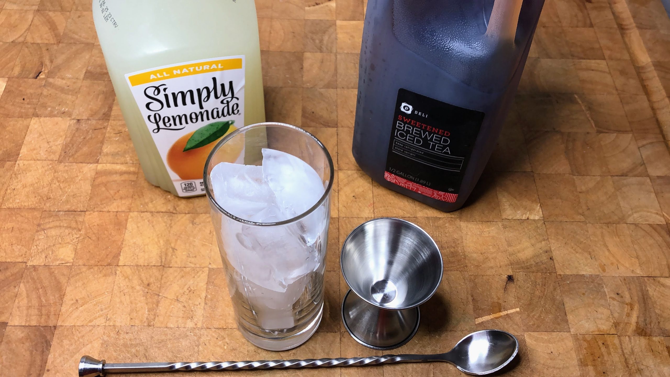 Highball glass with ice next to jigger, spoon, lemonade and iced tea.