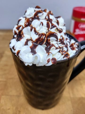 Biscoff hot chocolate in a mug topped with whipped cream and chocolate syrup and a container of biscoff spread in the background.