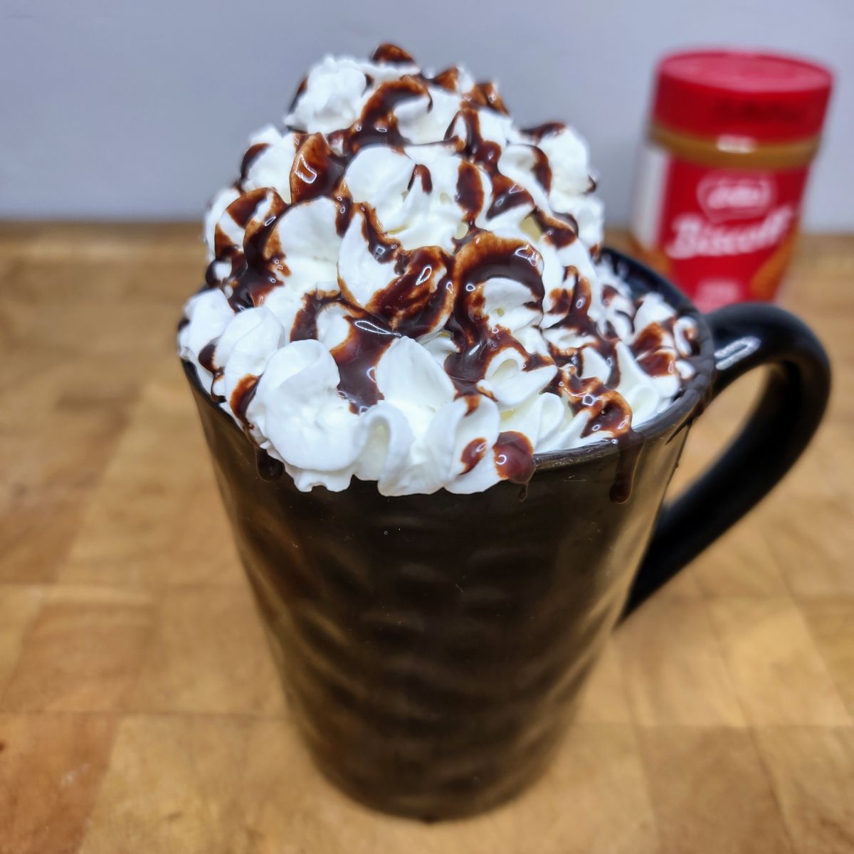 Biscoff hot chocolate in a mug topped with whipped cream and chocolate syrup and a container of biscoff spread in the background.