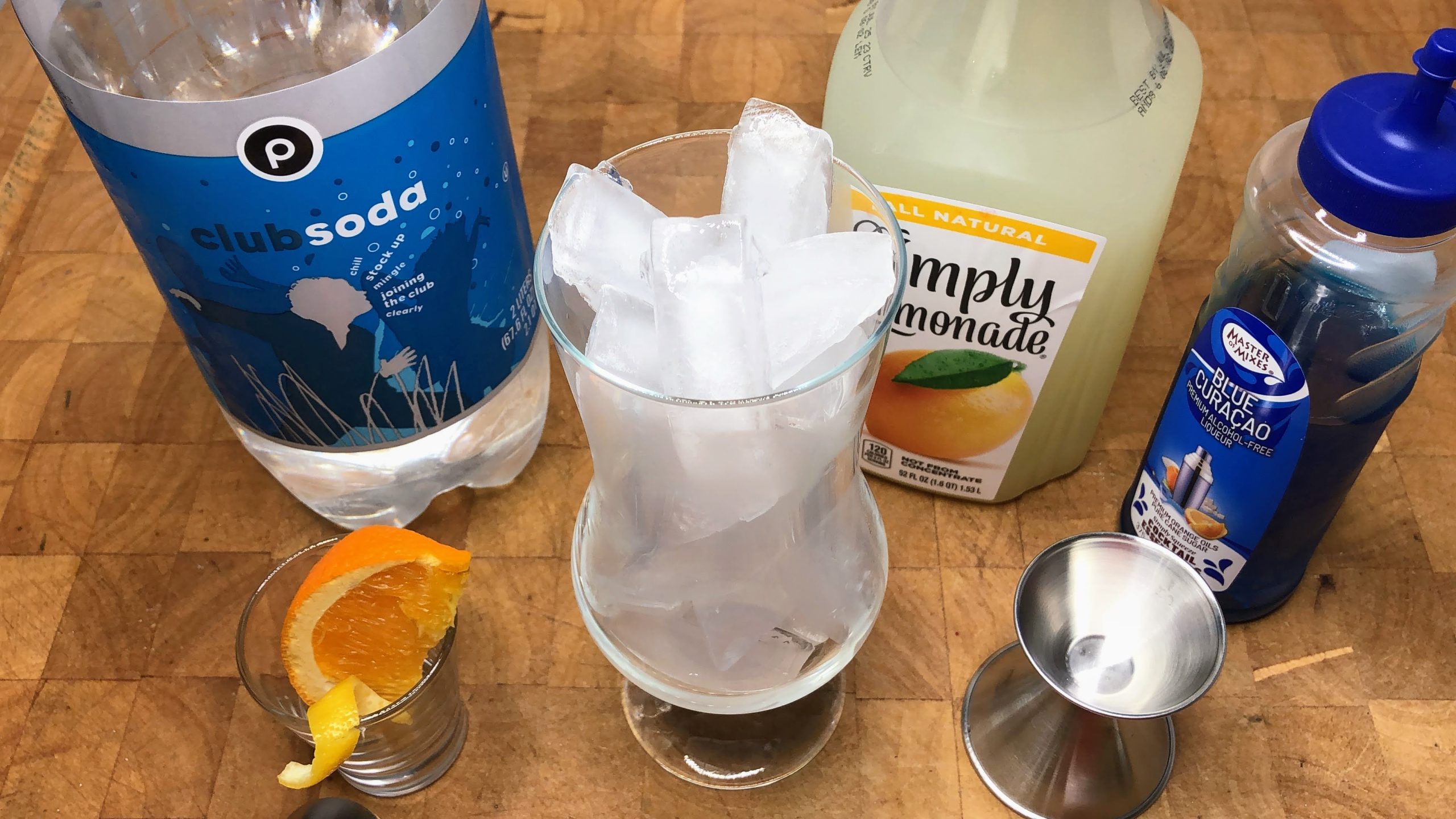 Hurricane glass with ice next to orange wedge, jigger, blue curaco, lemonade and club soda.