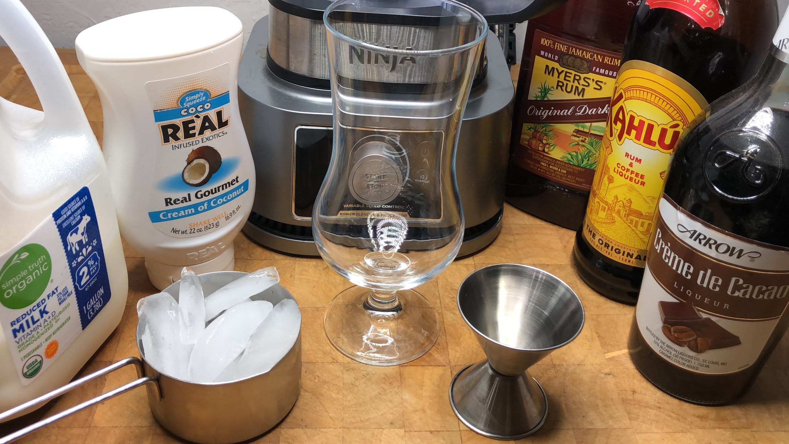 Ingredients for a bushwacker drink next to a cup of ice, hurricane glass, jigger and blender.
