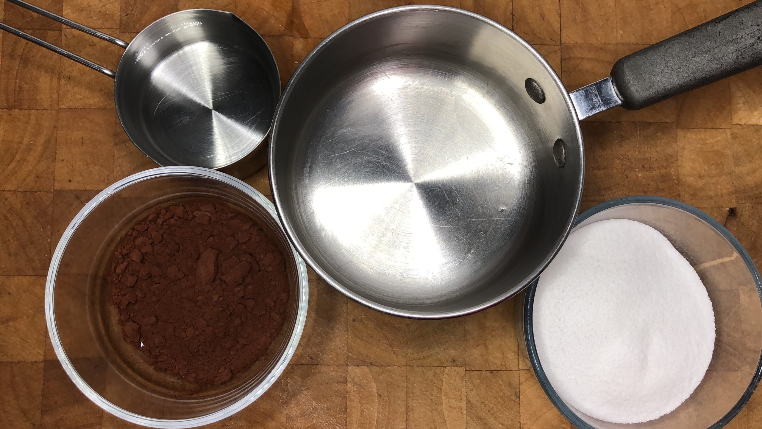 Bowls of chocolate, sugar and water next to a saucepan.