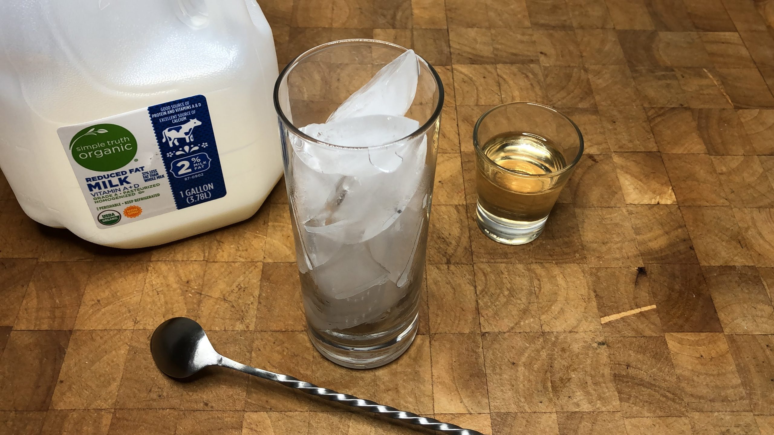 Highball glass with ice next to a bar spoon, cinnamon syrup and a carton of milk.