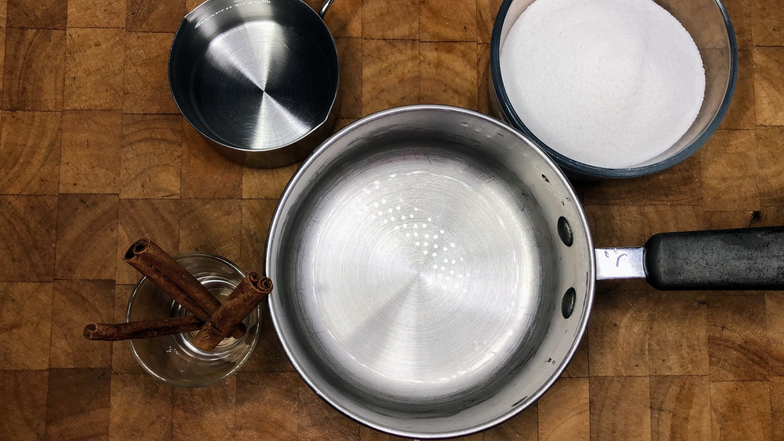 Bowls of cinnamon sticks, sugar and water next to a saucepan.