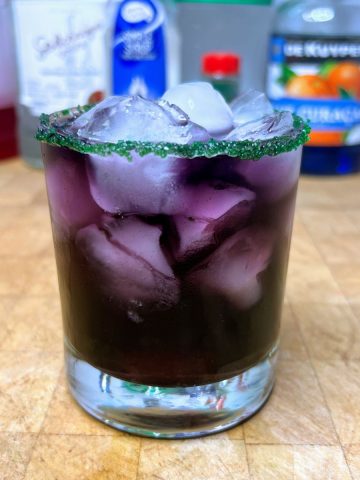 Drunk witch drink in a rocks glass with green sugar rim and ingredients in the background.