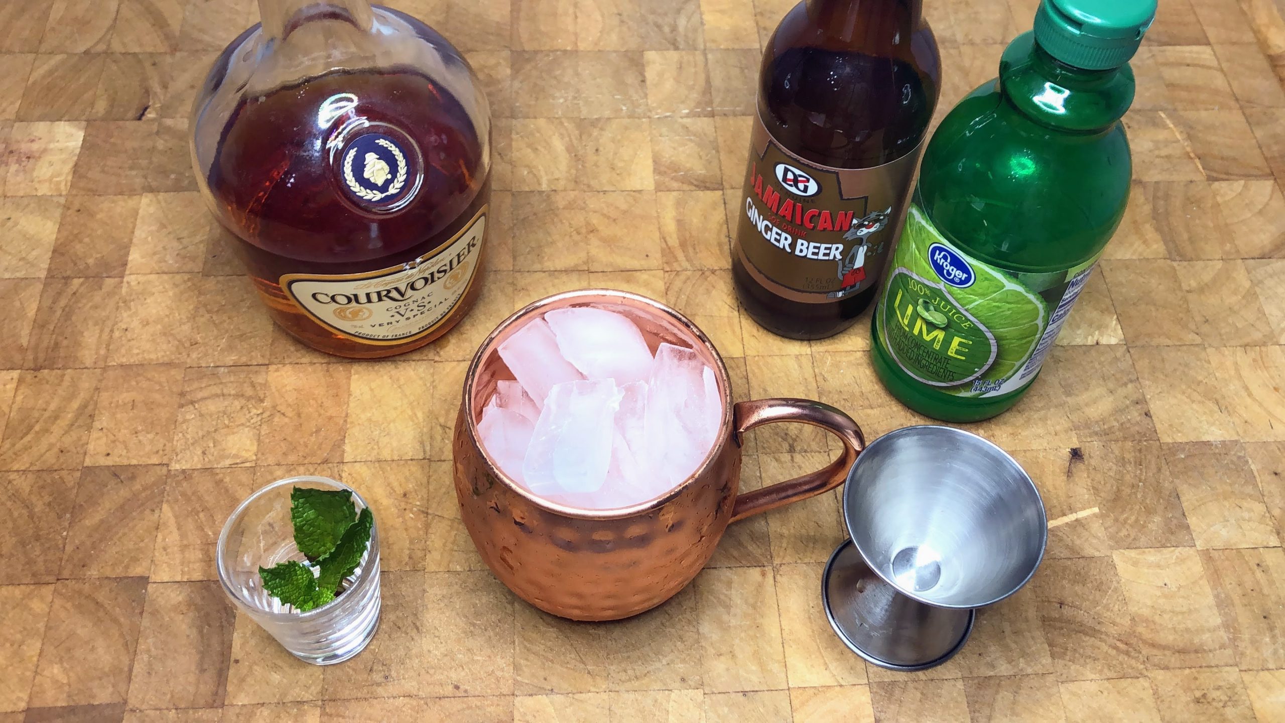 Copper mug filled with ice next to a jigger, mint leaves and bottles of cognac, ginger beer and lime juice.