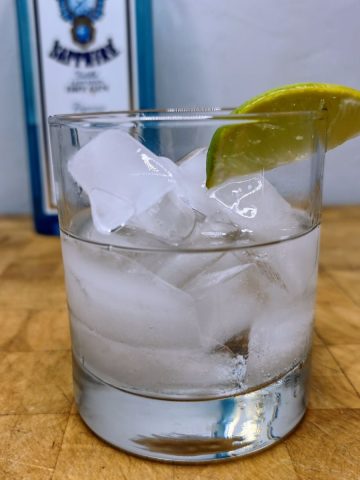 Gin on the rocks with a lime wedge on a wooden table with bottle of gin in the background.