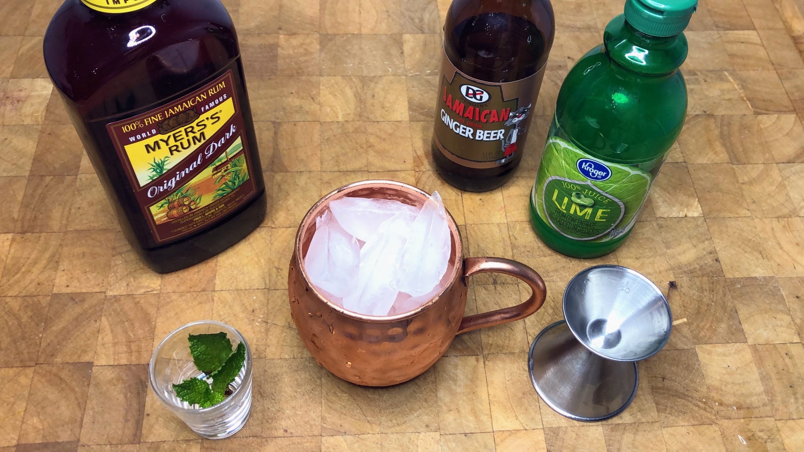 Copper mug with ice in it next to a jigger, mint leaves and bottles of jamaican rum, ginger beer and lime juice.
