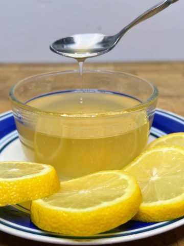Lemon simple syrup in a bowl on a saucer with lemon wheels next to it and the syrup being scooped by a spoon.
