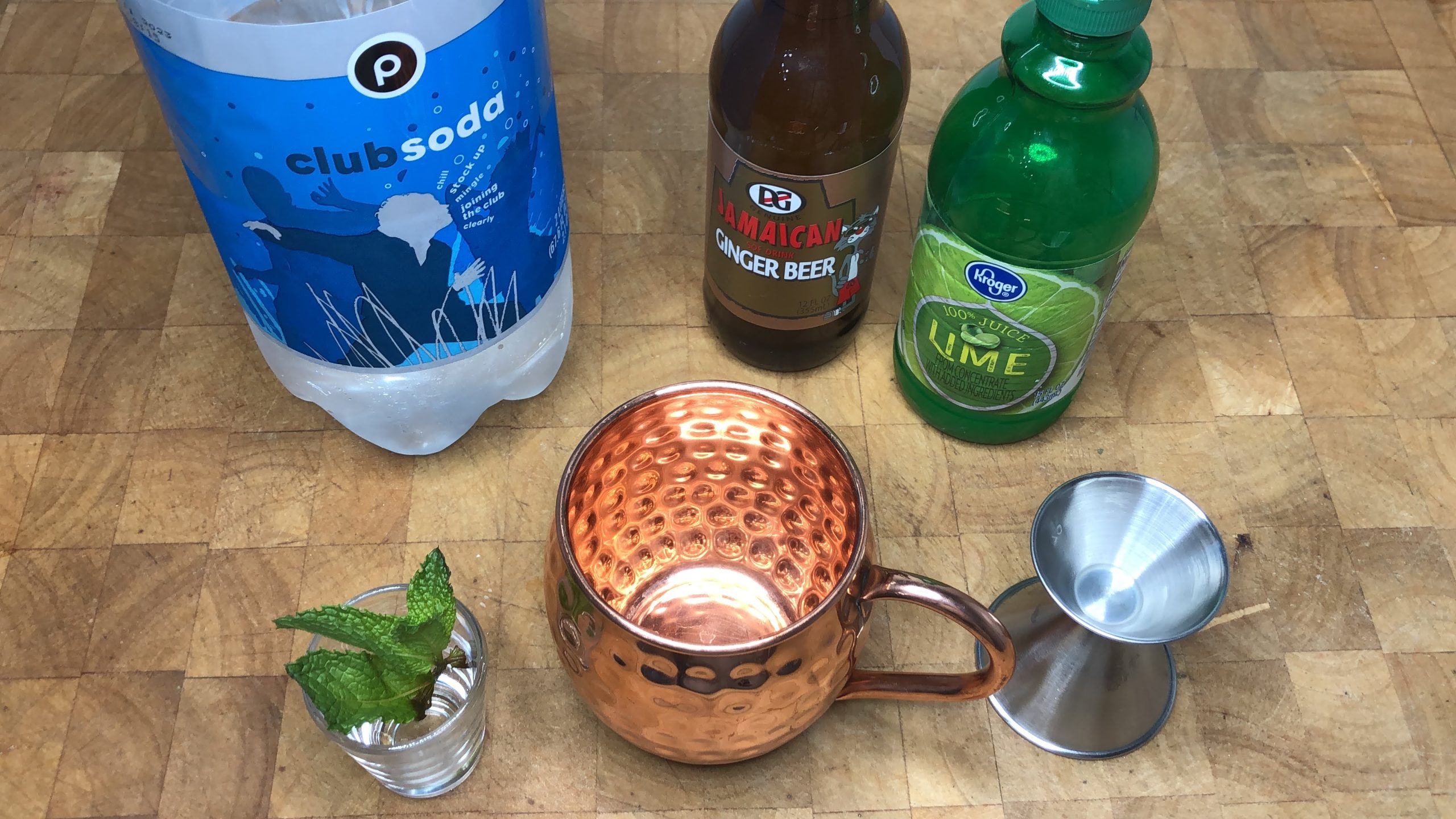 Copper mug, jigger and mint leaves in front of bottles of club soda, ginger beer and lime juice.