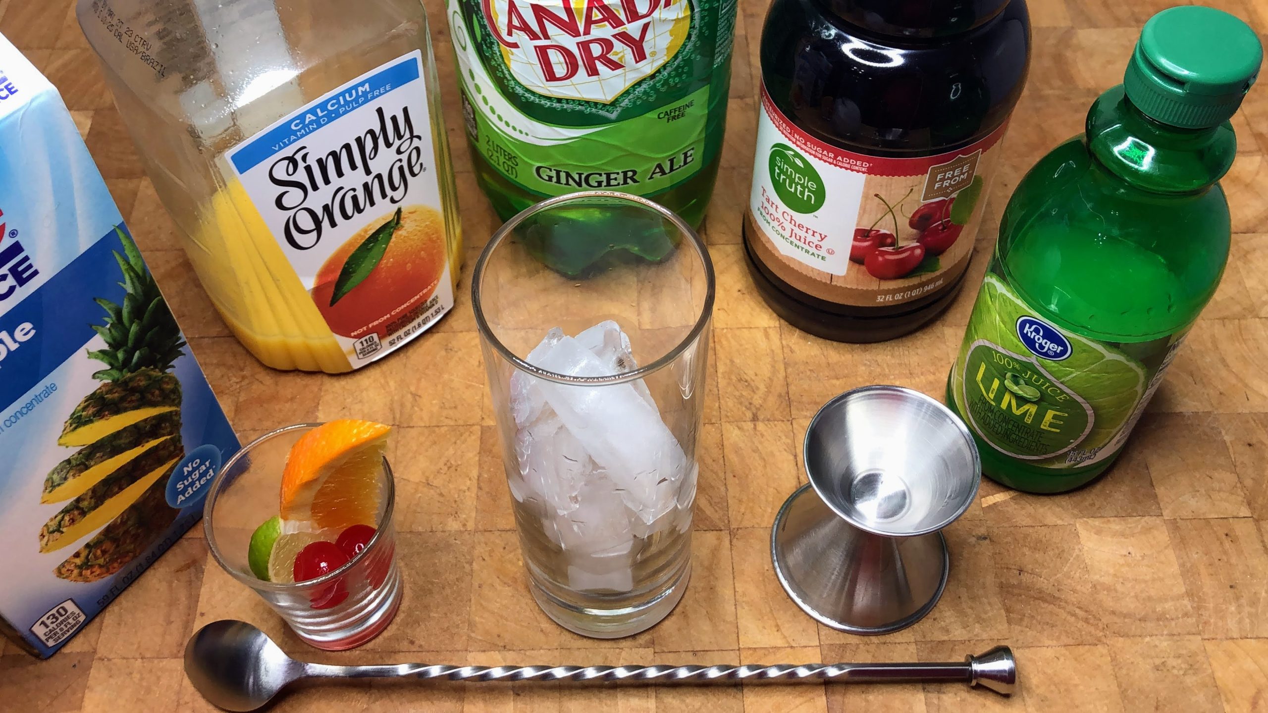 Pineapple, orange, cherry and lime juice next to ginger ale, highball glass, sliced fruit, jigger and bar spoon.