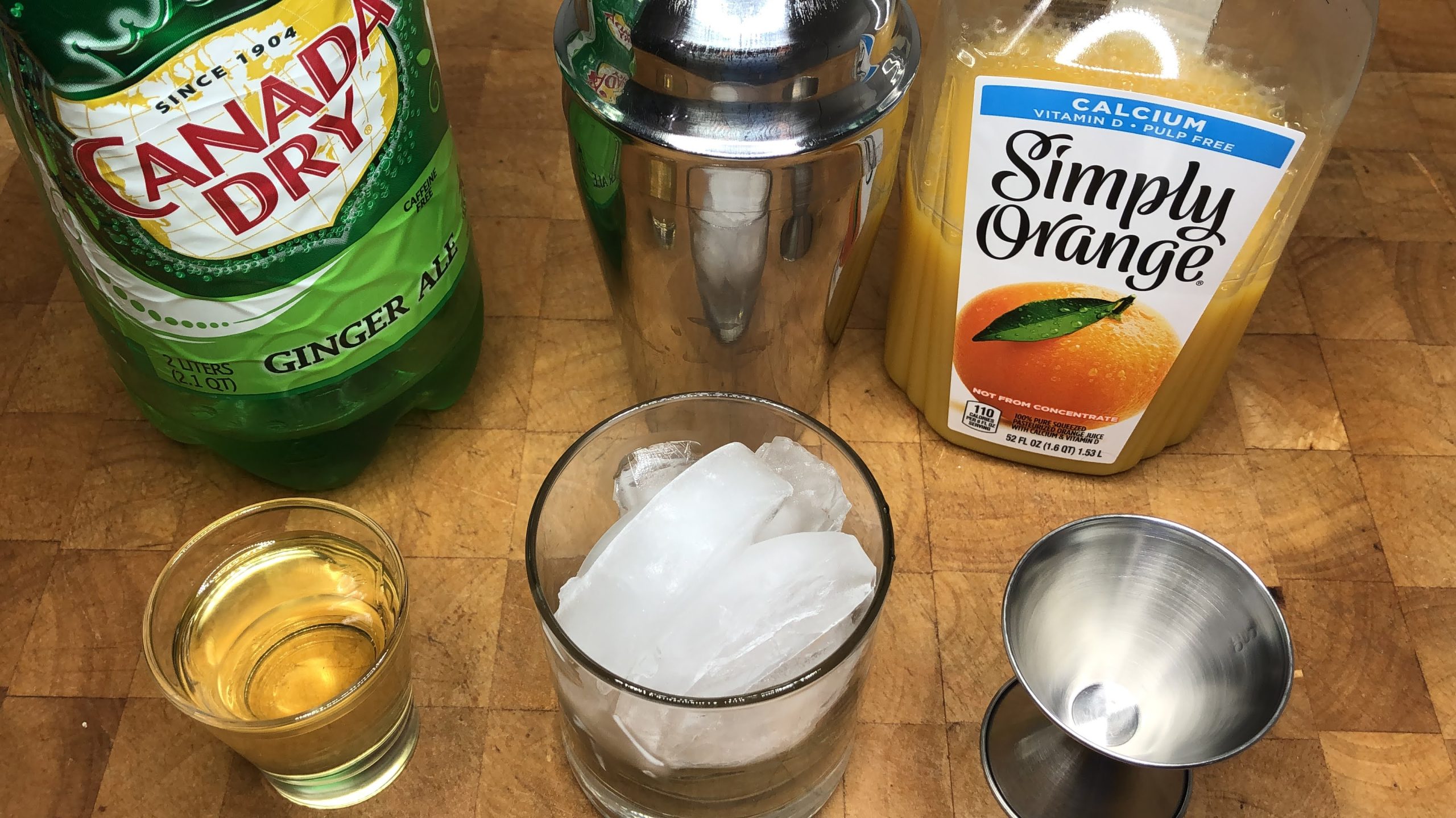 Rocks glass with ice next to orange simple syrup, a jigger, a shaker, orange juice and ginger ale.