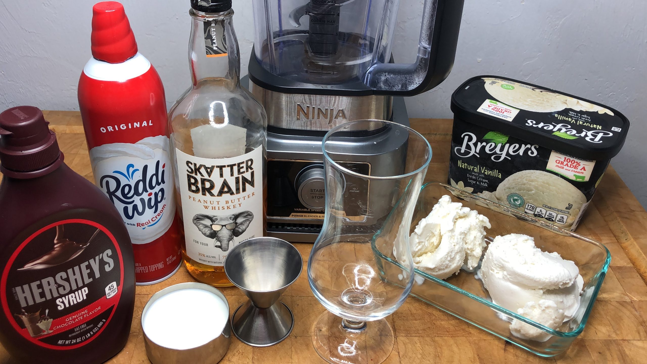 Chocolate syrup, whipped cream, milk and vanilla ice cream next to blender, jigger and hurricane glass.