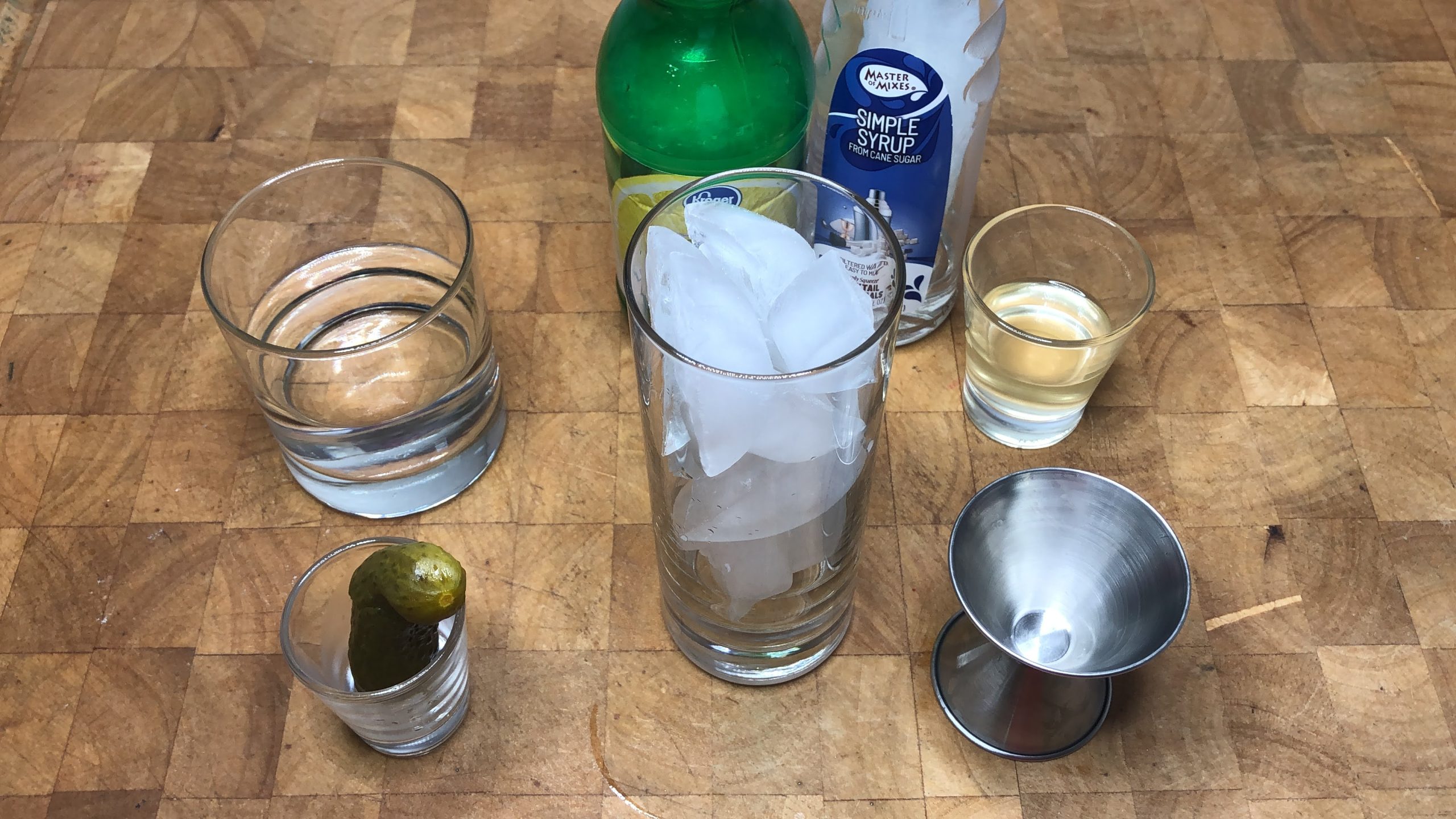 Highball glass with ice next to a pickle, jigger, water, lemon juice and simple syrup.