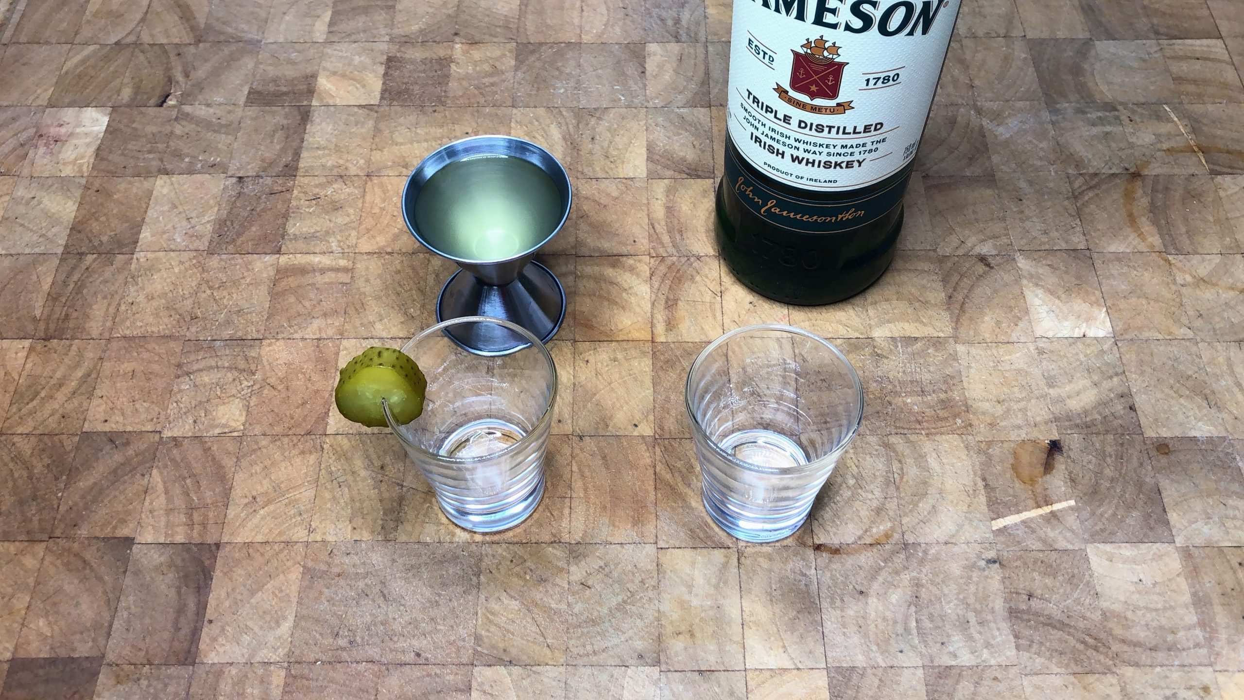 Whiskey next to a jigger with pickle juice and two shot glasses.