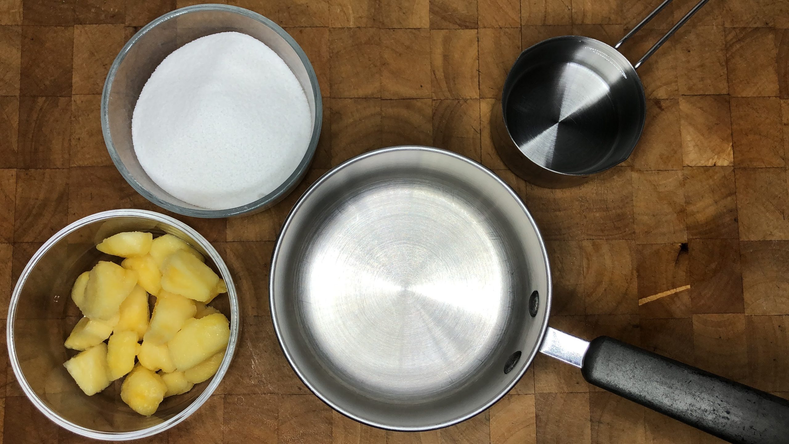 An empty pot next to bowls of pineapple, sugar and water.