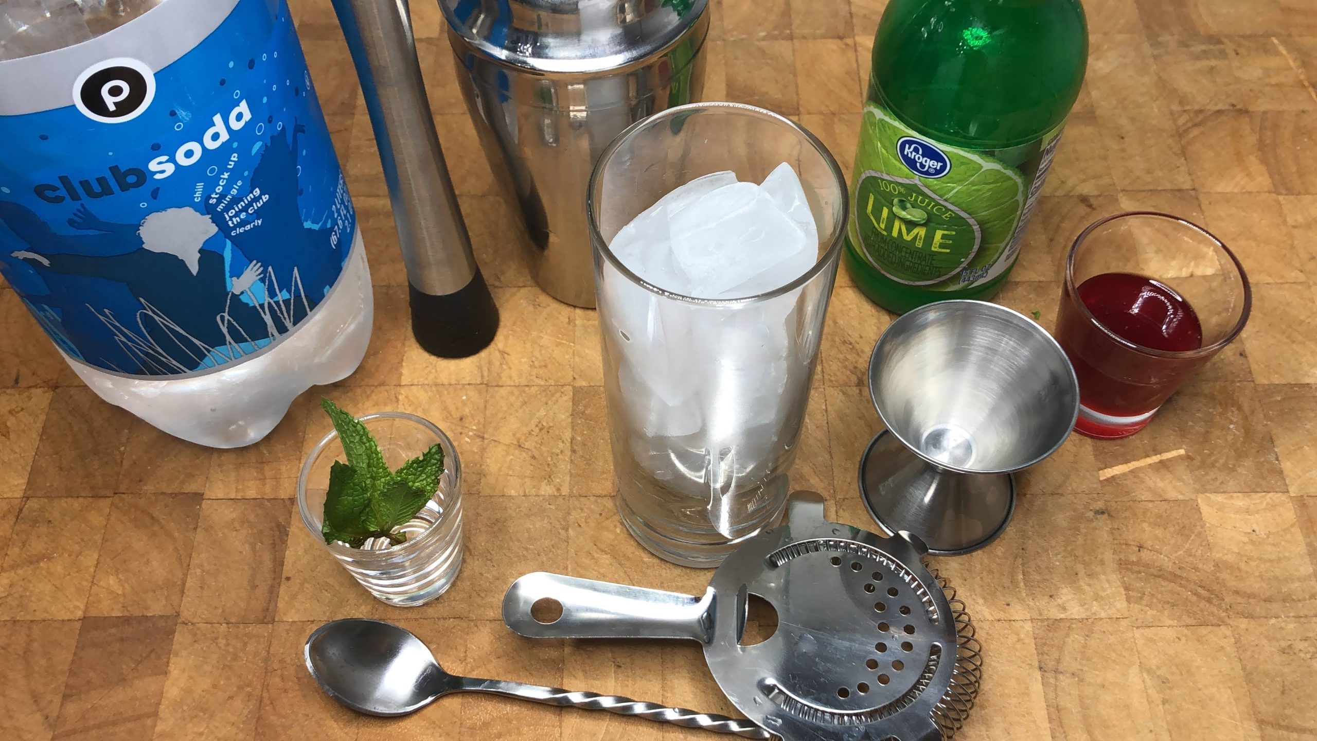 Highball glass with ice next to a jigger, strainer, spoon, muddler, shaker, mint leaves, raspberry syrup, lime juice and club soda.