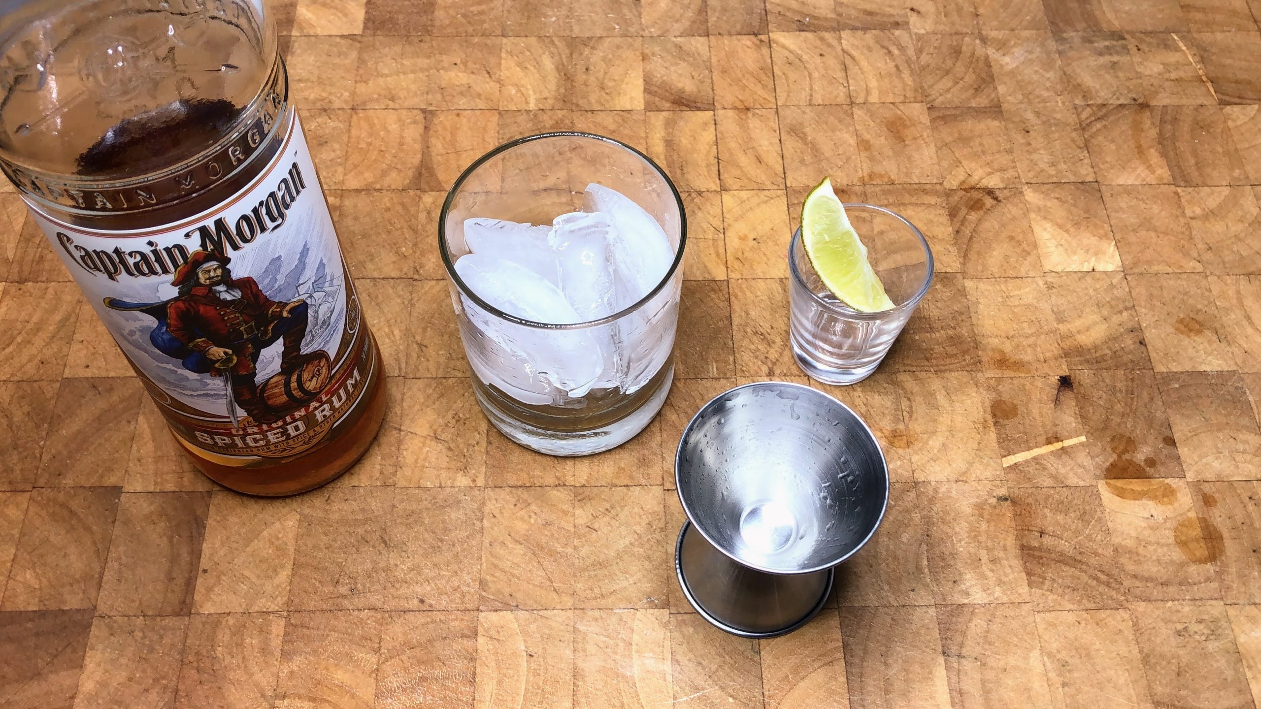 Bottle of spiced rum next to rocks glass with ice, lime wedge and a jigger.