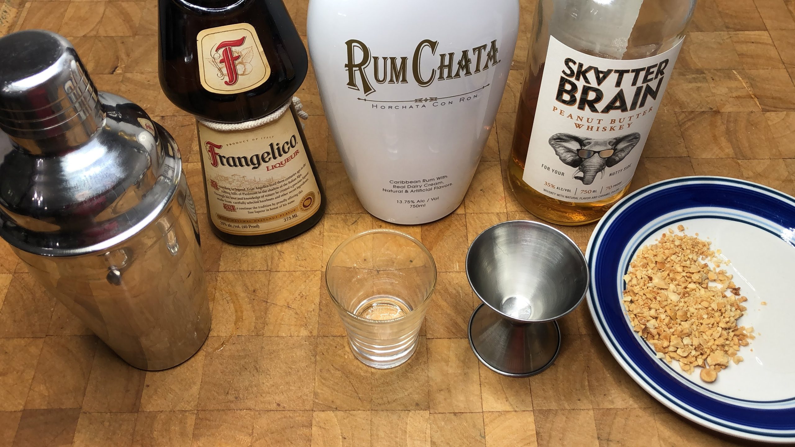 Frangelico, rumchata and peanut butter whiskey next to shot glass, jigger, shaker and saucer with crushed peanuts.