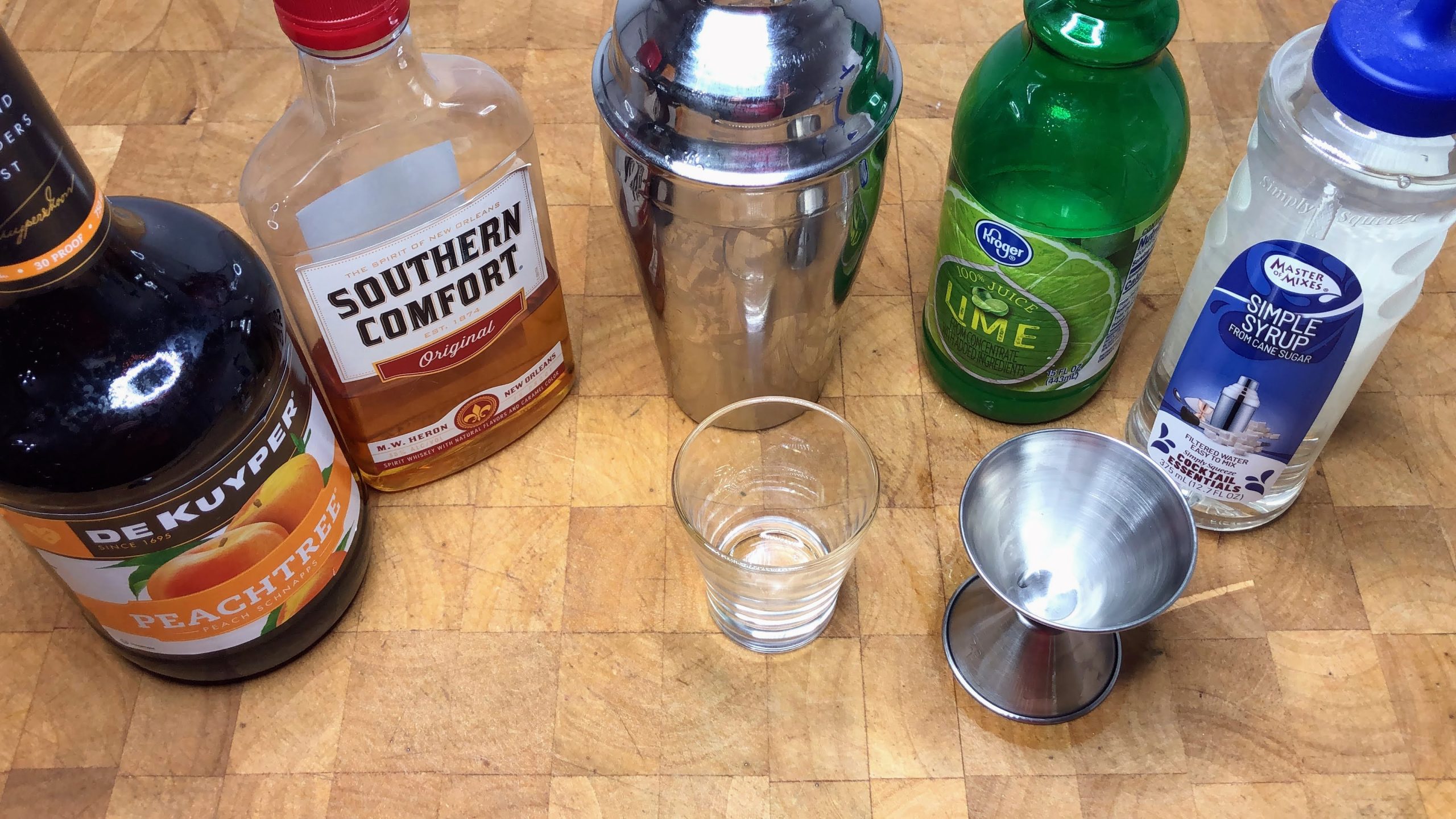 Peach schnapps, southern comfort, lime juice and simple syrup next to shot glass, cocktail shaker and jigger.