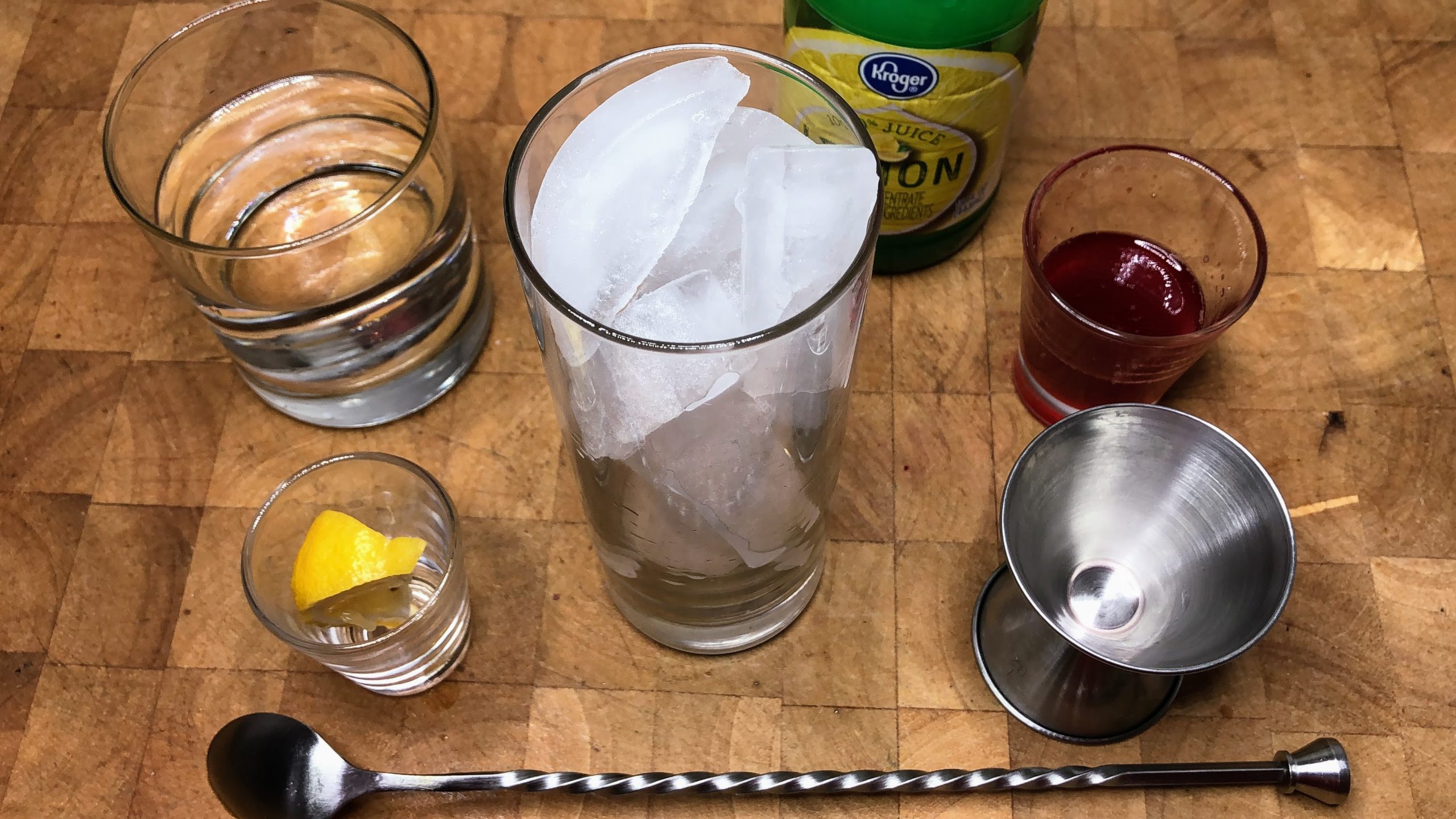 Highball glass with ice next to water, jigger, lemon juice, strawberry simple syrup and a bar spoon.