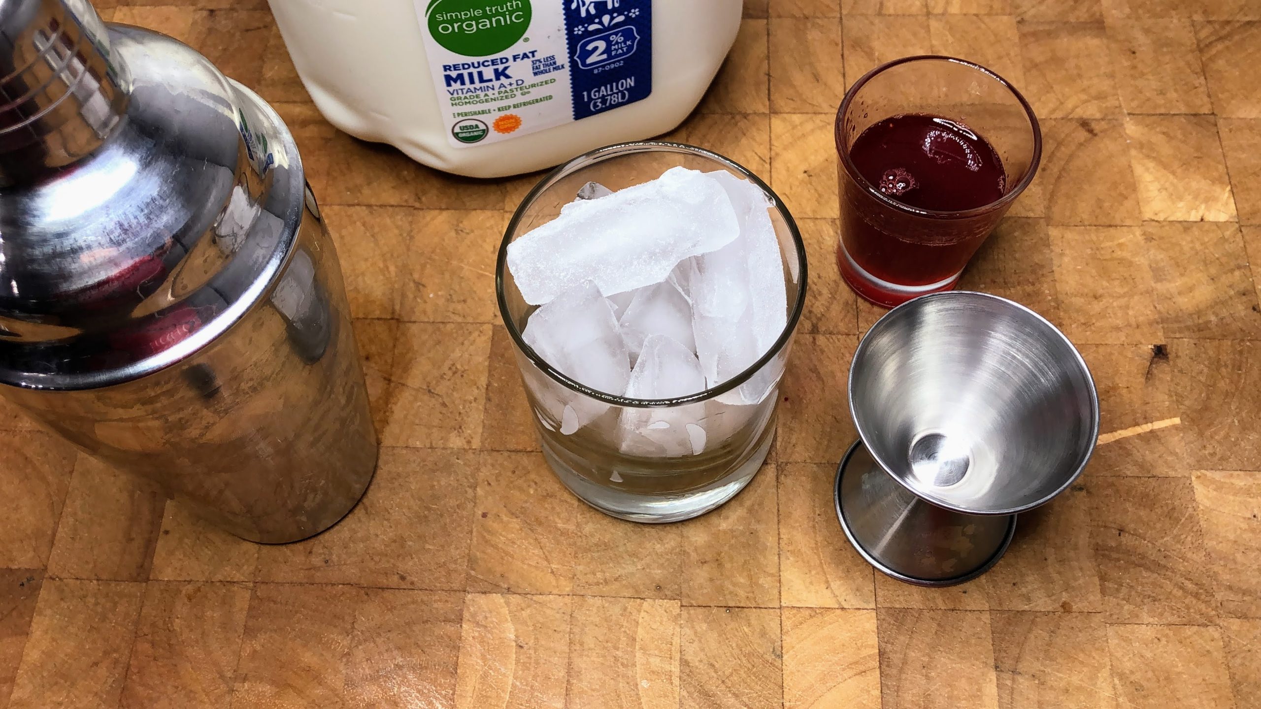 Shaker and rocks glass next to jigger, strawberry simple syrup and milk.