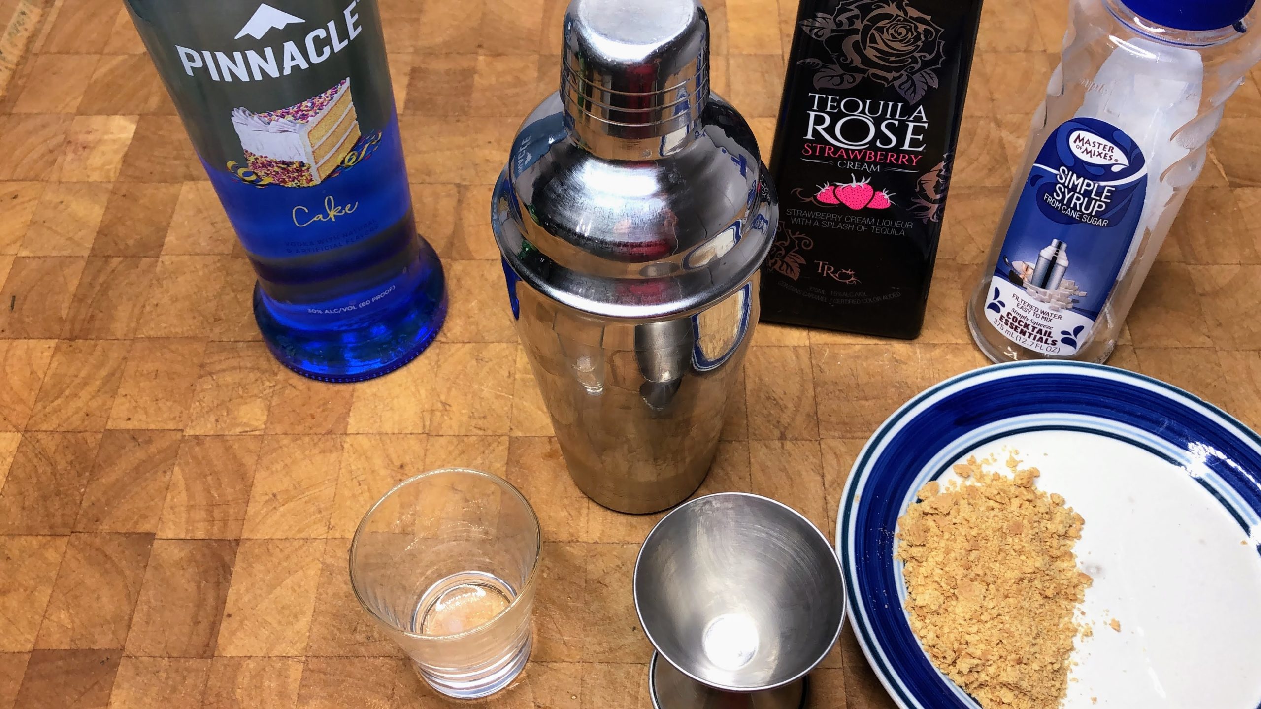Cake vodka, tequila rose and simple syrup next to a shaker, shot glass, jigger and saucer with crumbled graham crackers.