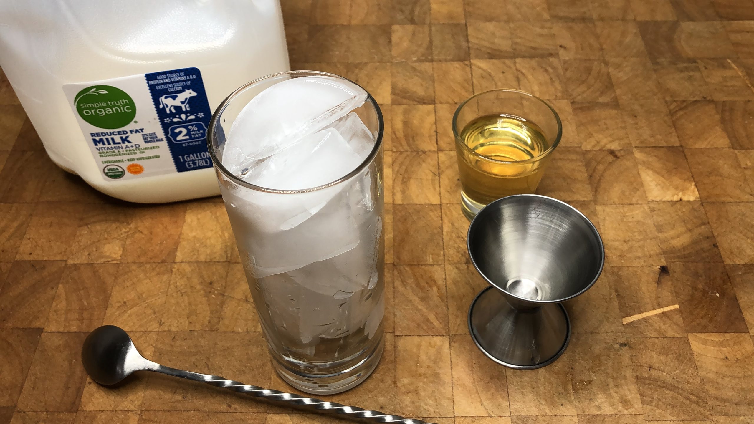 Milk and sugar cookie syrup next to highball glass with ice, jigger and bar spoon.