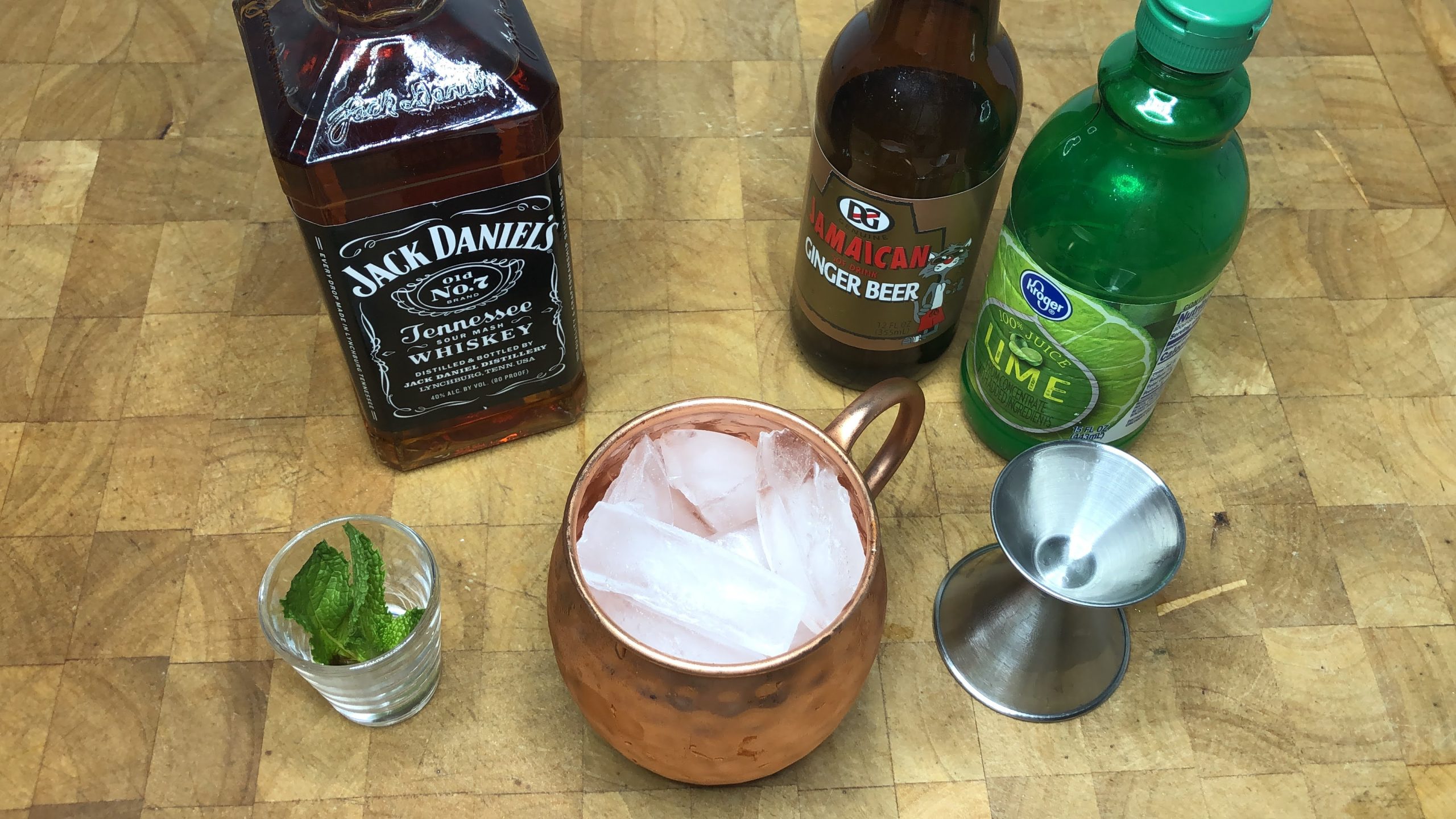 Mug with ice next to jigger, lime juice, ginger beer and jack daniels.