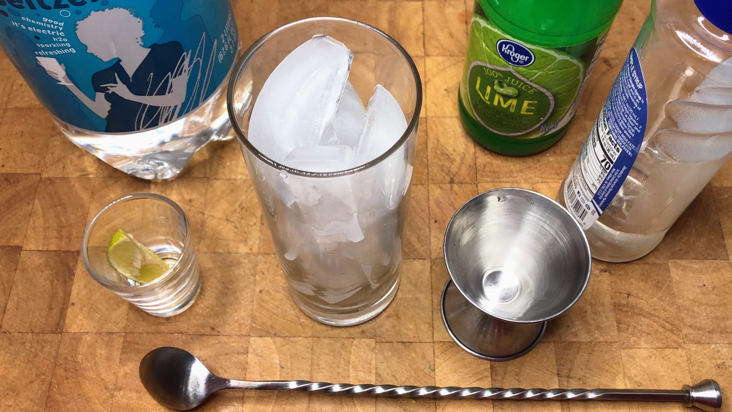 Highball glass with ice next to spoon, jigger, simple syrup, lime juice and seltzer water.