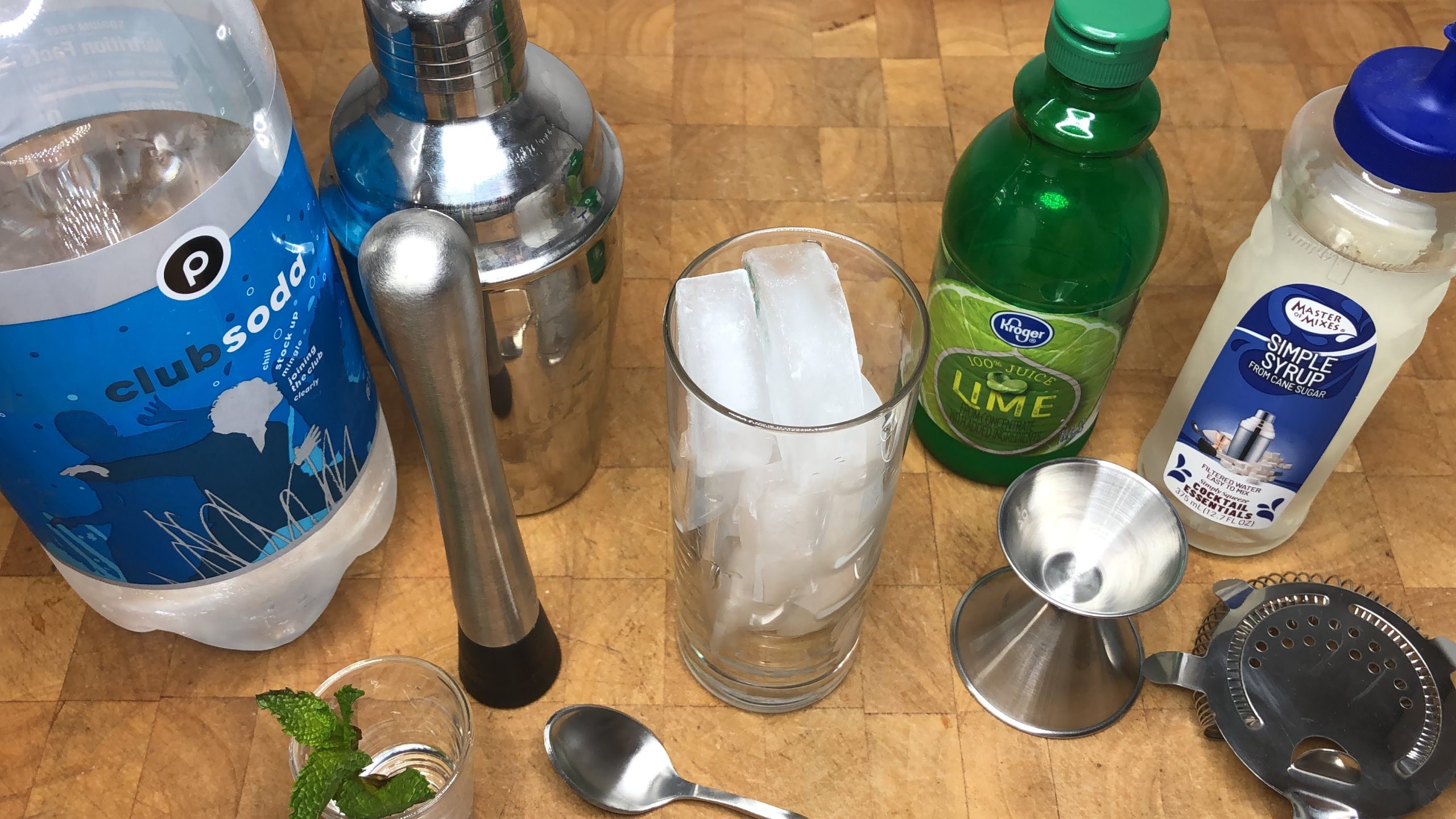 Club soda, lime juice, mint leaves and simple syrup next to a muddler, spoon, shaker, glass, and jigger.