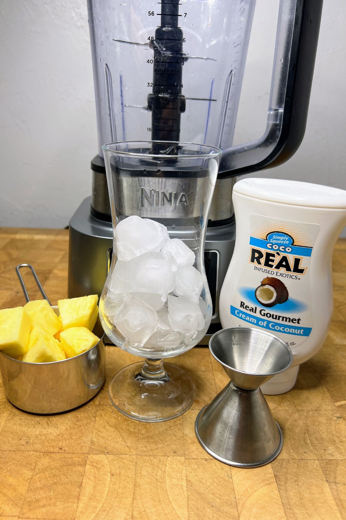 Pineapple, hurricane glass with ice, jigger and cream of coconut in front of a blender.