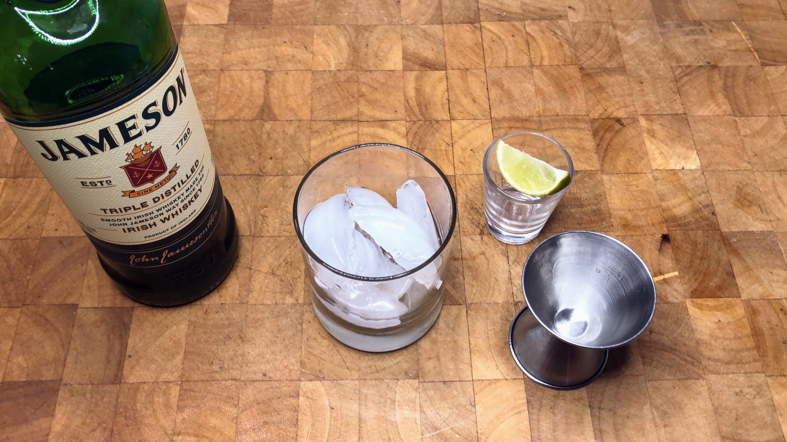 Whiskey, rocks glass with ice, lime wedge and a jigger on a wooden table.