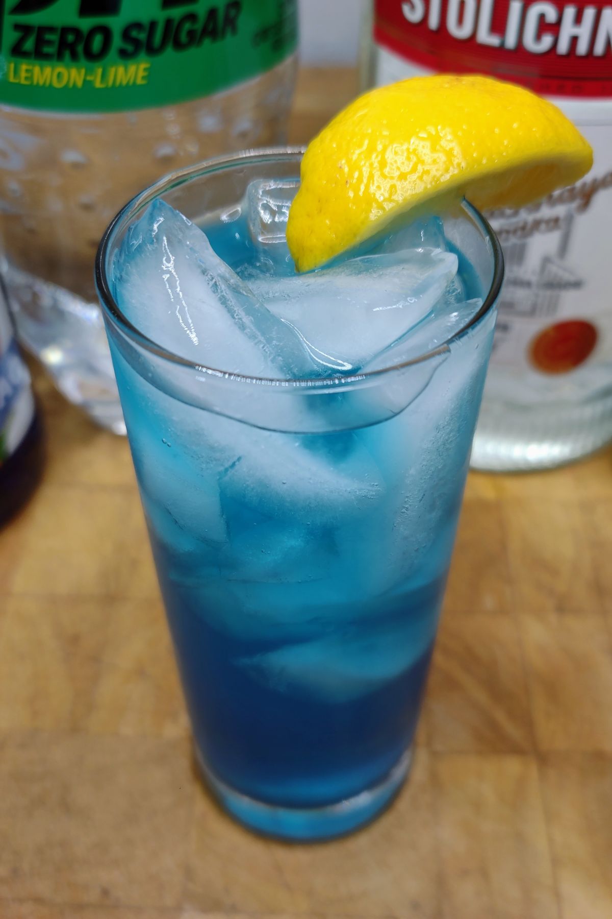 Electric lemonade cocktail on a wooden table with ingredients behind the glass.