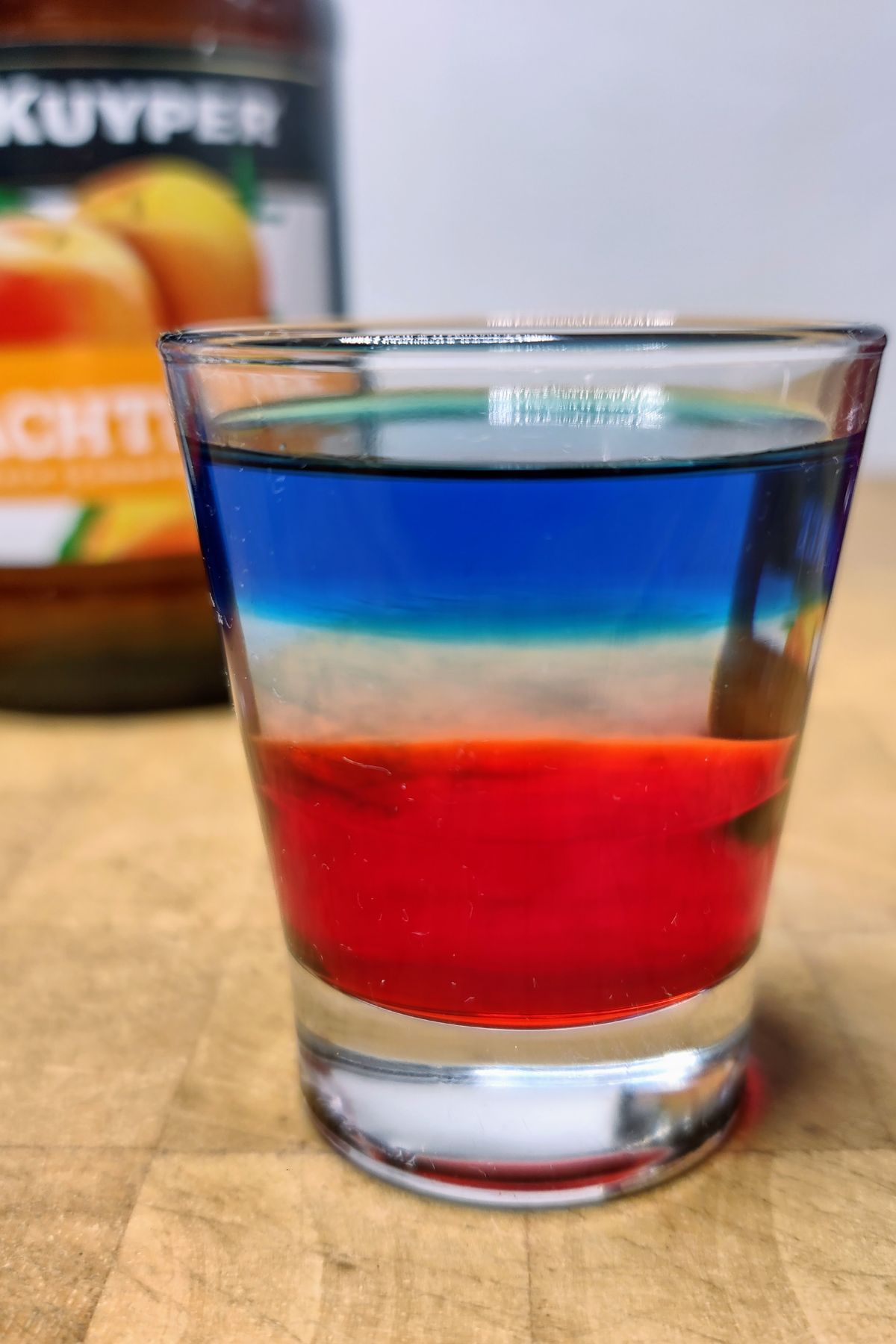 Red white and blue shot on a wooden table with ingredients behind the glass.